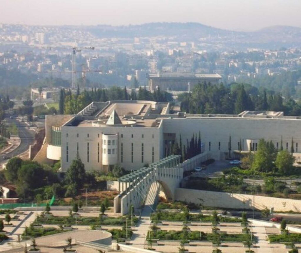 ein großer modernerer Gebäudekomplex mit einer Grünanlage davor. Im Hintergrund sieht man eine Stadt