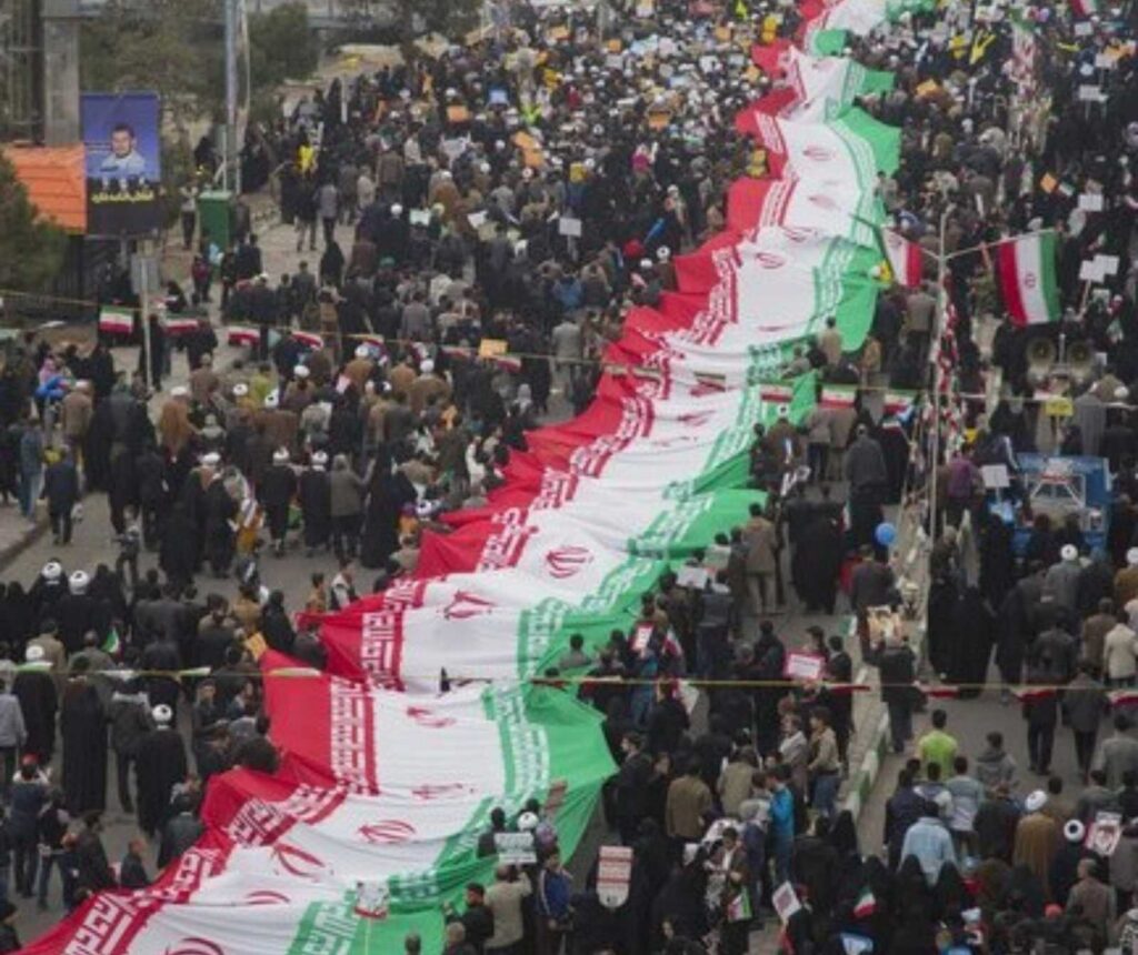 ein großer Menschenauflauf auf einer Straße aus der Luft aufgenommen. In der Mitte veerläuft eine lange Stoffbahn mit der Abbildung der iranischen Flagge