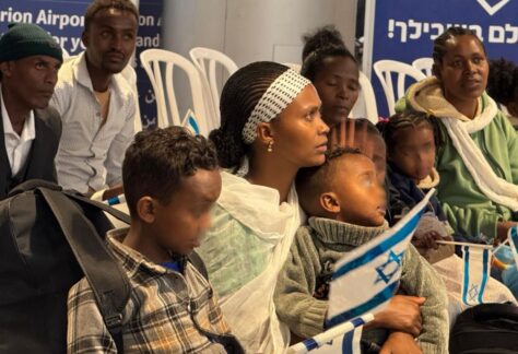 mehrere dunkelhäutige Menschen, Frauen, Kinder und Männer, sitzen auf weißen Plastigstühlen in Reihen hintereinander. Manche halten israelische Fähnchen in der Hand