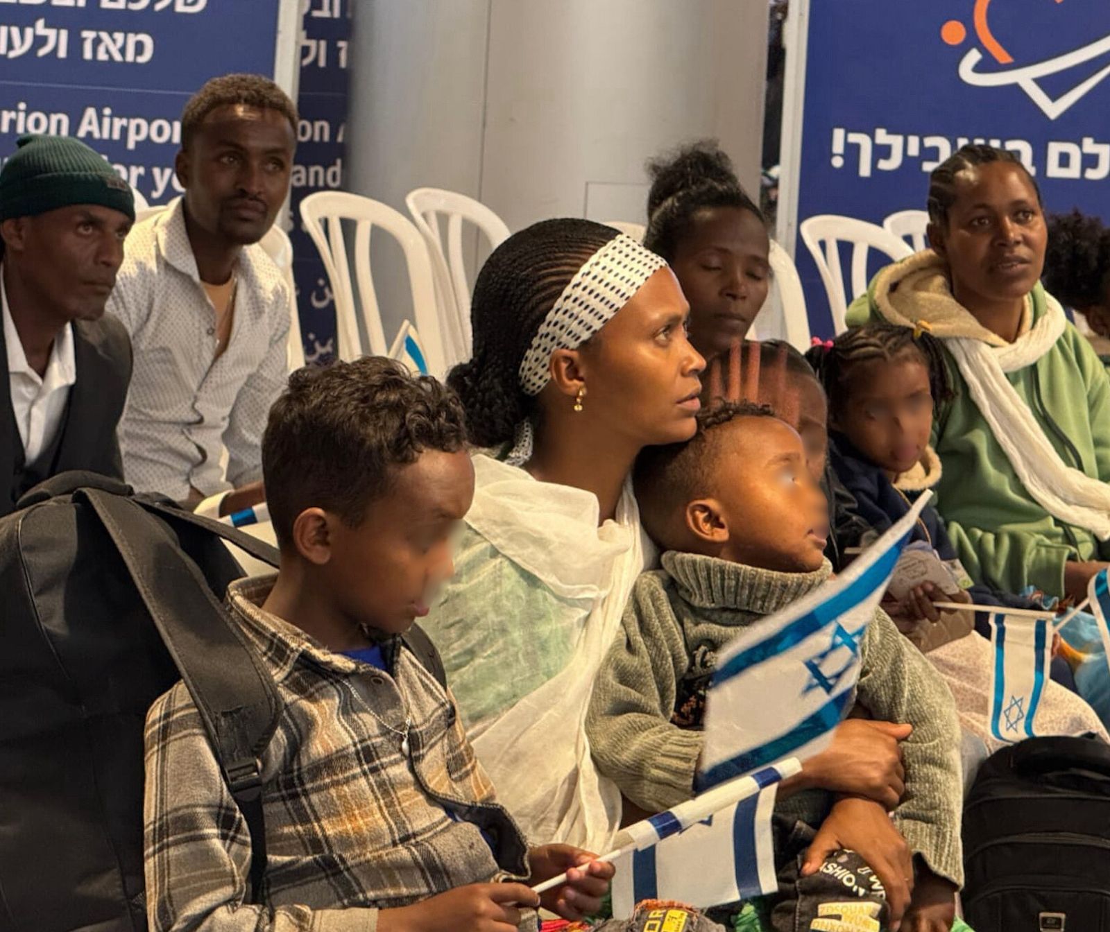 mehrere dunkelhäutige Menschen, Frauen, Kinder und Männer, sitzen auf weißen Plastigstühlen in Reihen hintereinander. Manche halten israelische Fähnchen in der Hand