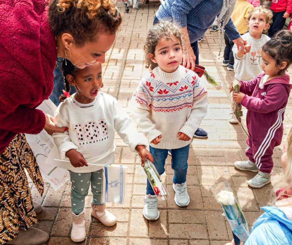 eine Frau beugt sich zu mehreren Kleinkindern hinunter. Eines hält ein Israelfähnchen in der Hand