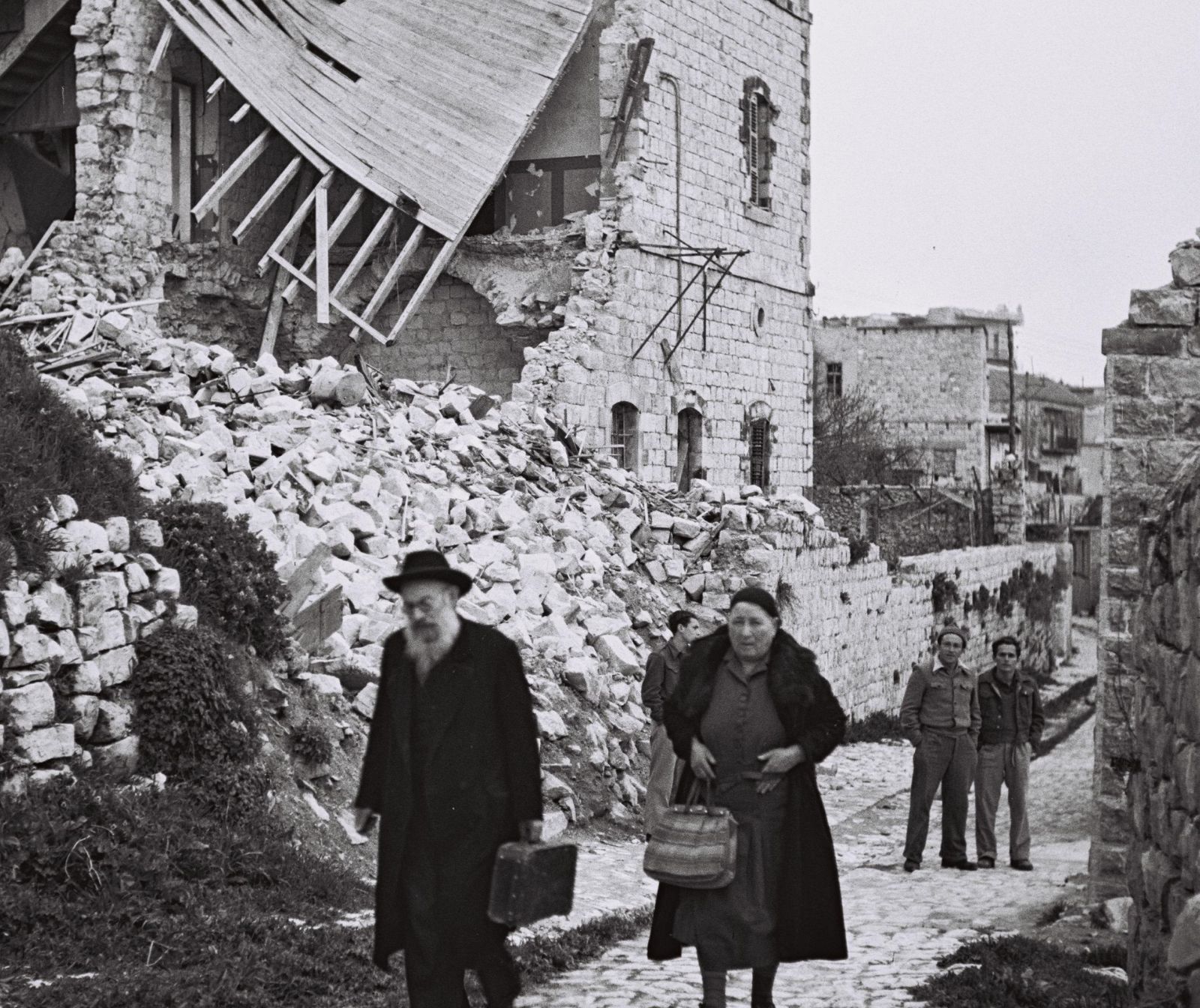 schwarz weiß Foto zerstörter Häuser. Menschen laufen mit Taschen die Straße entlang