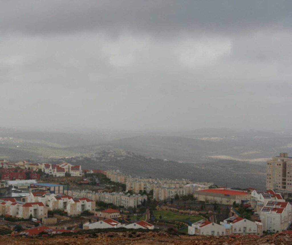 Weitblick über eine Stadt mit weißen Häusern und roten Dächern auf hügeligem Gelände. Der Himmel ist dunstig