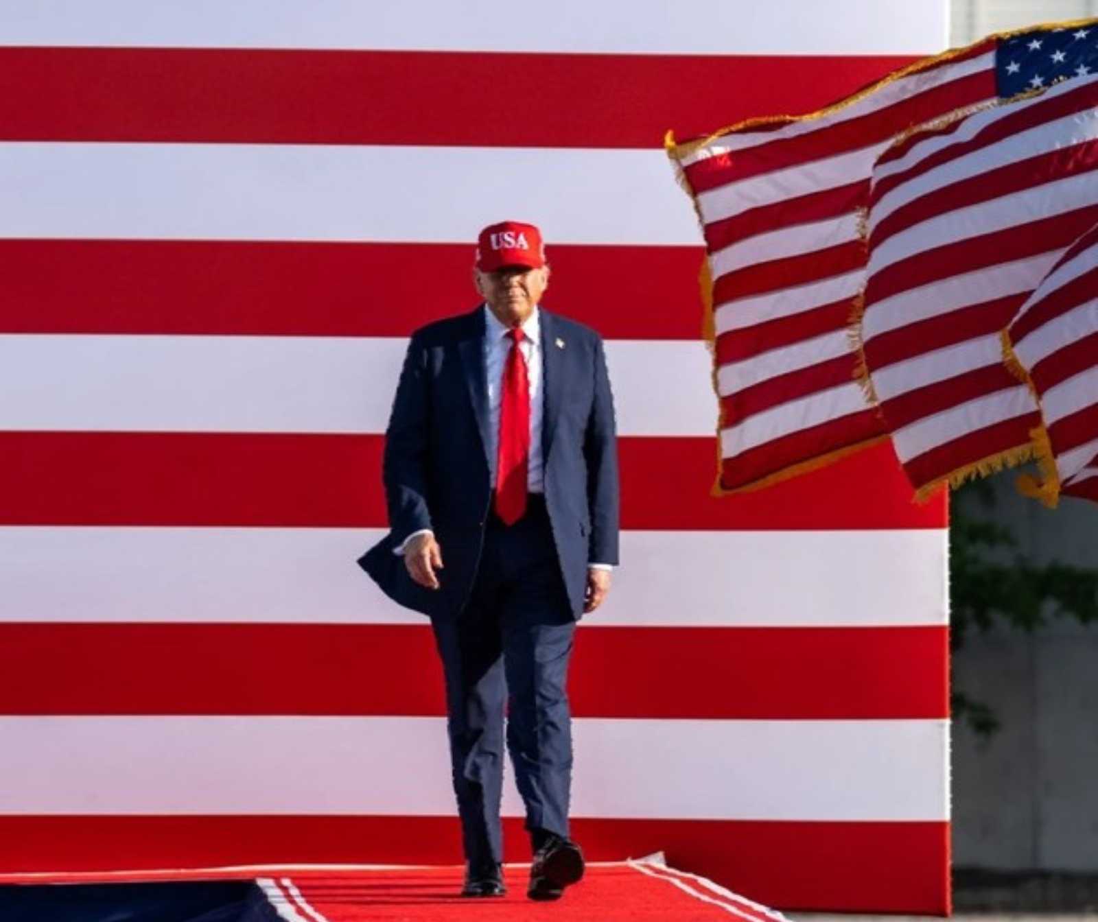 Ein Mann im blauem Anzug, roter Krawatte und roter Baseballmütze laufen über einen roten Teppich. Backdrop sind große weiß-rote Querstreifen, es wehen links amerikanische Flaggen