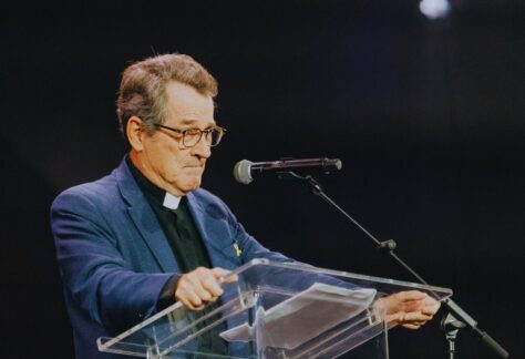 ein Mann an einem Rednerpult aus plexiglas und einem Mikrofon. Er trägt eine Brille und Anzug