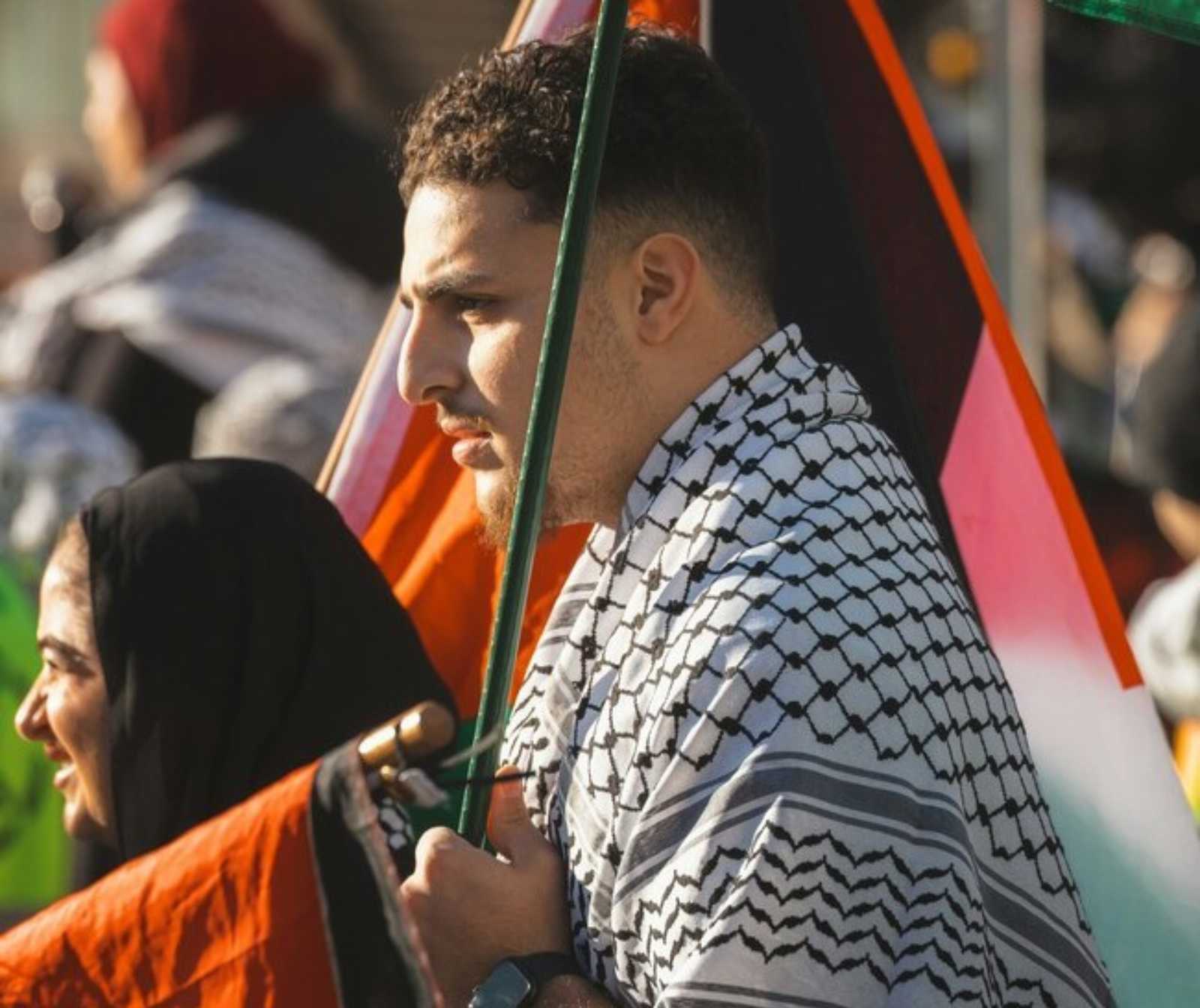 ein junger Mann mit kurzem, lockigem Haar trägt ein schwarz-weiß gemustertes Tuch um die Schulter.Er hält eine Stange; im Hintergrund weitere Menschen mit Fahnen