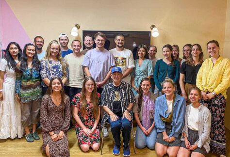 ein alter Mann auf einem Stuhl sitzend ist umringt von einer Gruppe von junger Erwachsener. Sie stehen oder sind kniend auf dem Boden; alle Blicken in die Kamera