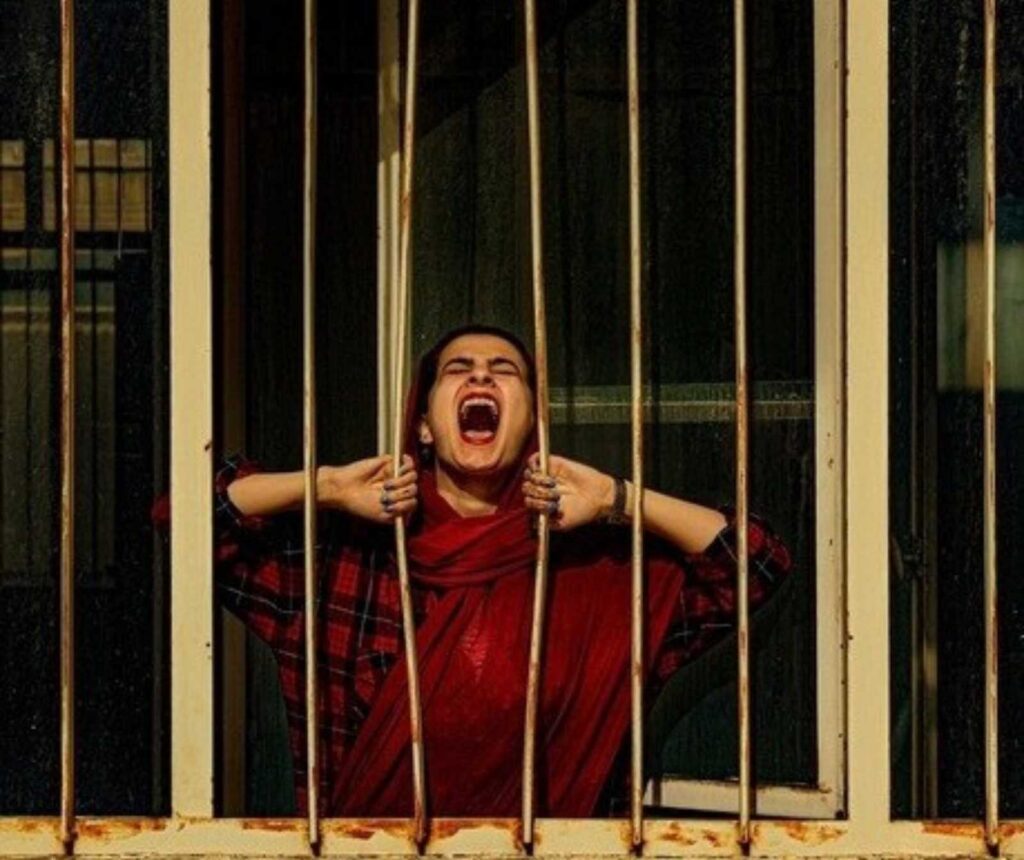 eine Frau in rotem Gewand hinter Gitterstäben. Sie hält zwei, die etwas auseinanderstehen, in der Hand und schreit durch diese hindurch