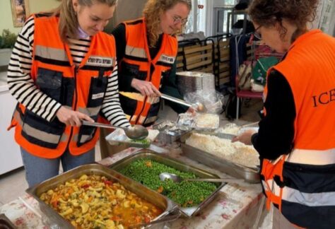 Frauem mit orangenfarbenen Warnwesten schöpfen Essen in Aluminiumschalen. Sie wirken fröhlich.