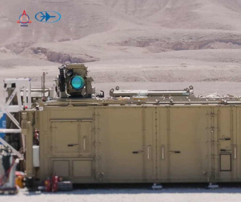 ein sandfarbener Container, auf dessen Dach ein Gerät mit runder blauer Linse montiert ist. Im Hintergrund ist eine Wüstenlandschaft zu sehen.