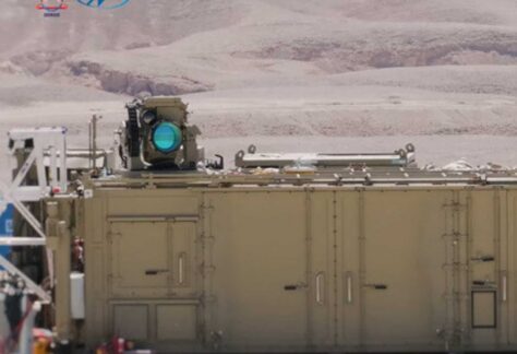 ein sandfarbener Container, auf dessen Dach ein Gerät mit runder blauer Linse montiert ist. Im Hintergrund ist eine Wüstenlandschaft zu sehen.