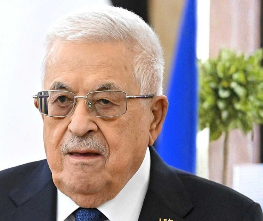 ein älterer Mann mit weißem Haar und Brille. Er trägt einen blauen Anzug, ein weißes Hemd mit blauer Krawatte