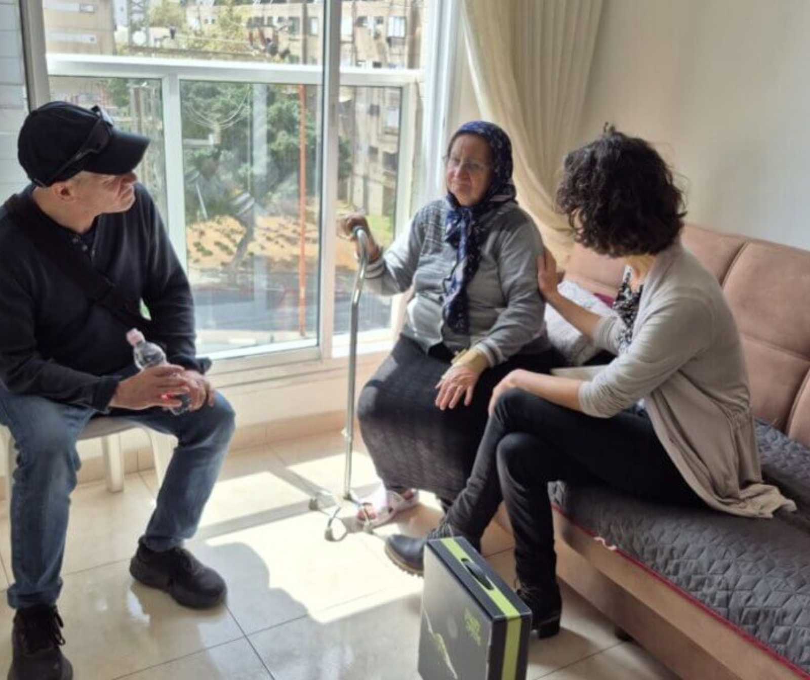 eine alte und eine jüngere Frau sitzen auf einem Sofa. Gegenüber ein Mann. Sie schauenzur alten Dame rüber