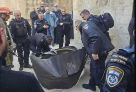 Mehrere Soicherheitskräfte und Einsatzpersonen in einer engen Straße begutachten ein großes beschädigtes Metallteilam Boden.