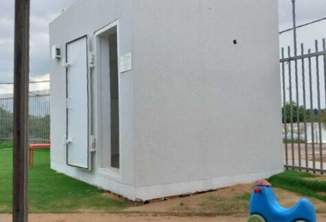 ein weißes rechteckiges containerartiges Häuschen. Die Tür steht offen, es ist ein Kleinkindfahrzeug zu sehen.