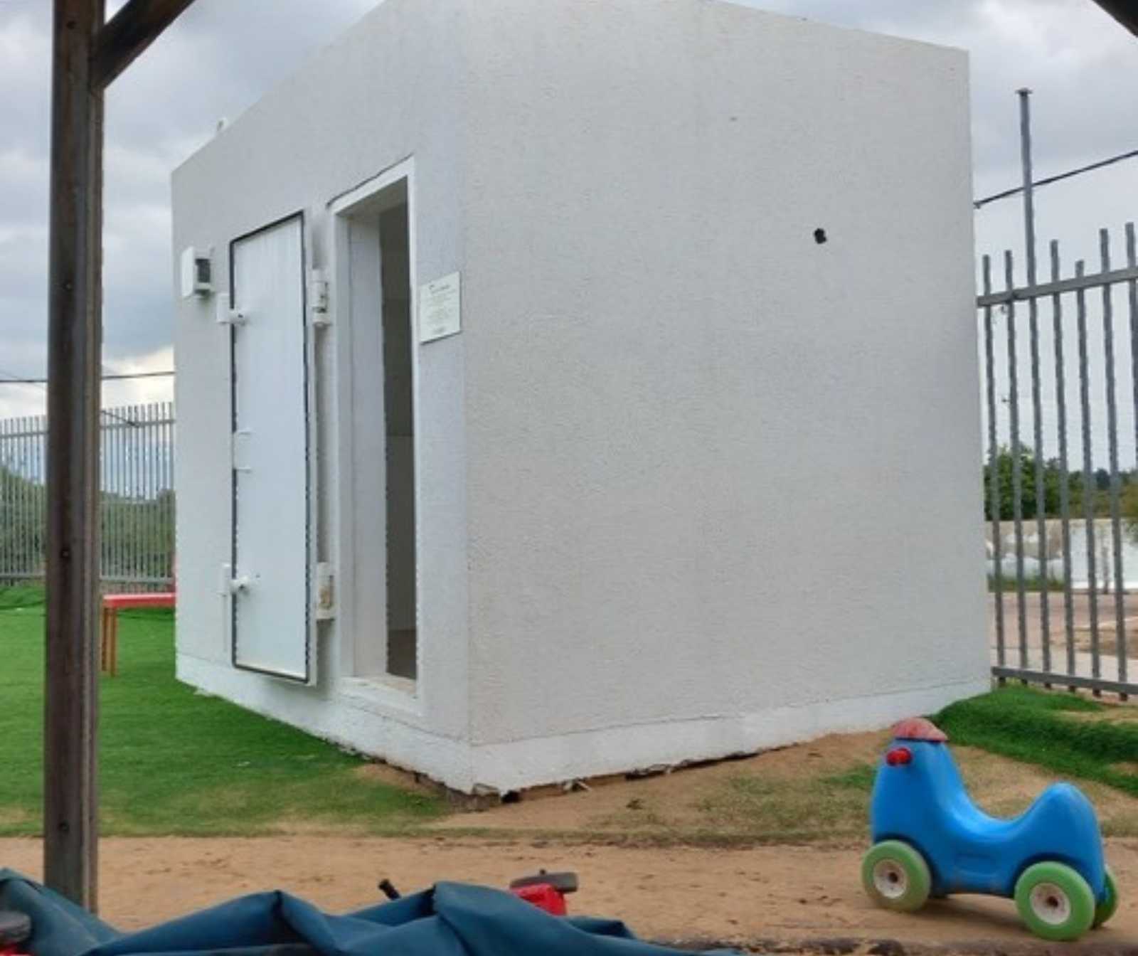 ein weißes rechteckiges containerartiges Häuschen. Die Tür steht offen, es ist ein Kleinkindfahrzeug zu sehen.