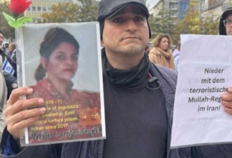 Ein Mann hält in seiner linken Hand ein Blatt mit der Aufschrift "Nieder mit dem terroristischen Mulah-Regime im Iran!" hoch. In der rechten hält er einen Ausdruck einer Abbildung einer Frau.