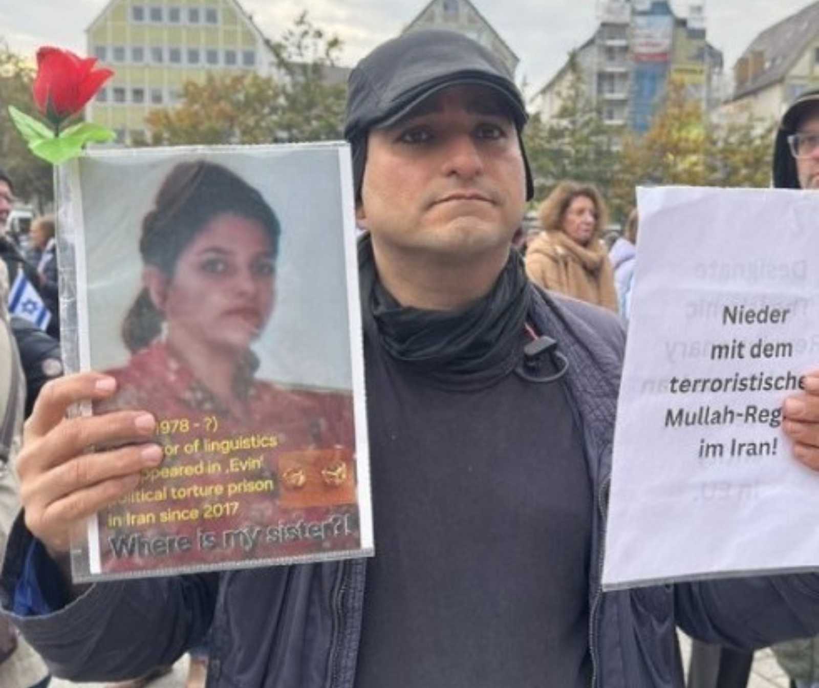 Ein Mann hält in seiner linken Hand ein Blatt mit der Aufschrift "Nieder mit dem terroristischen Mulah-Regime im Iran!" hoch. In der rechten hält er einen Ausdruck einer Abbildung einer Frau.