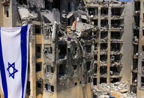 ein Zerstörter Wohnkomplex. Das Fensterglas ist nicht mehr vorhanden, Trümmer und Ruß, sowie eine israelische Fahne sind zu sehen.