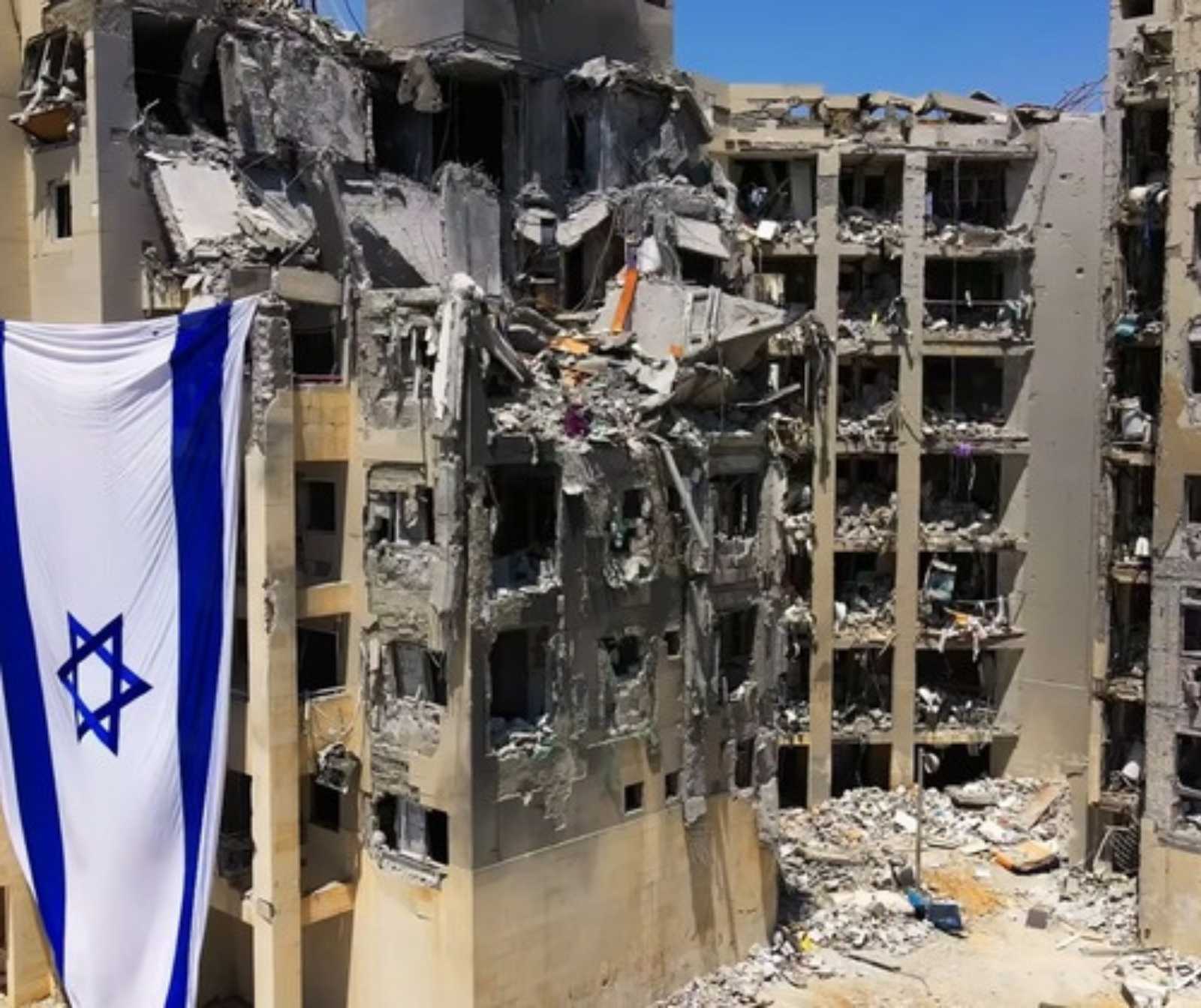 ein Zerstörter Wohnkomplex. Das Fensterglas ist nicht mehr vorhanden, Trümmer und Ruß, sowie eine israelische Fahne sind zu sehen.