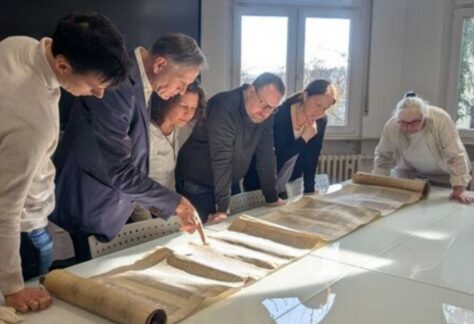 an einem langen Tisch stehen sechs Personen und schauen sich eine ausgebreitete Schriftrolle an