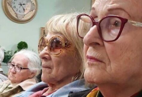 drei alte Damen nebeneinander schauen in die gleiche Richtung. An der Wand des Zimmers hängt eine Uhr; Pflanzen sind im Hintergrund zu sehen