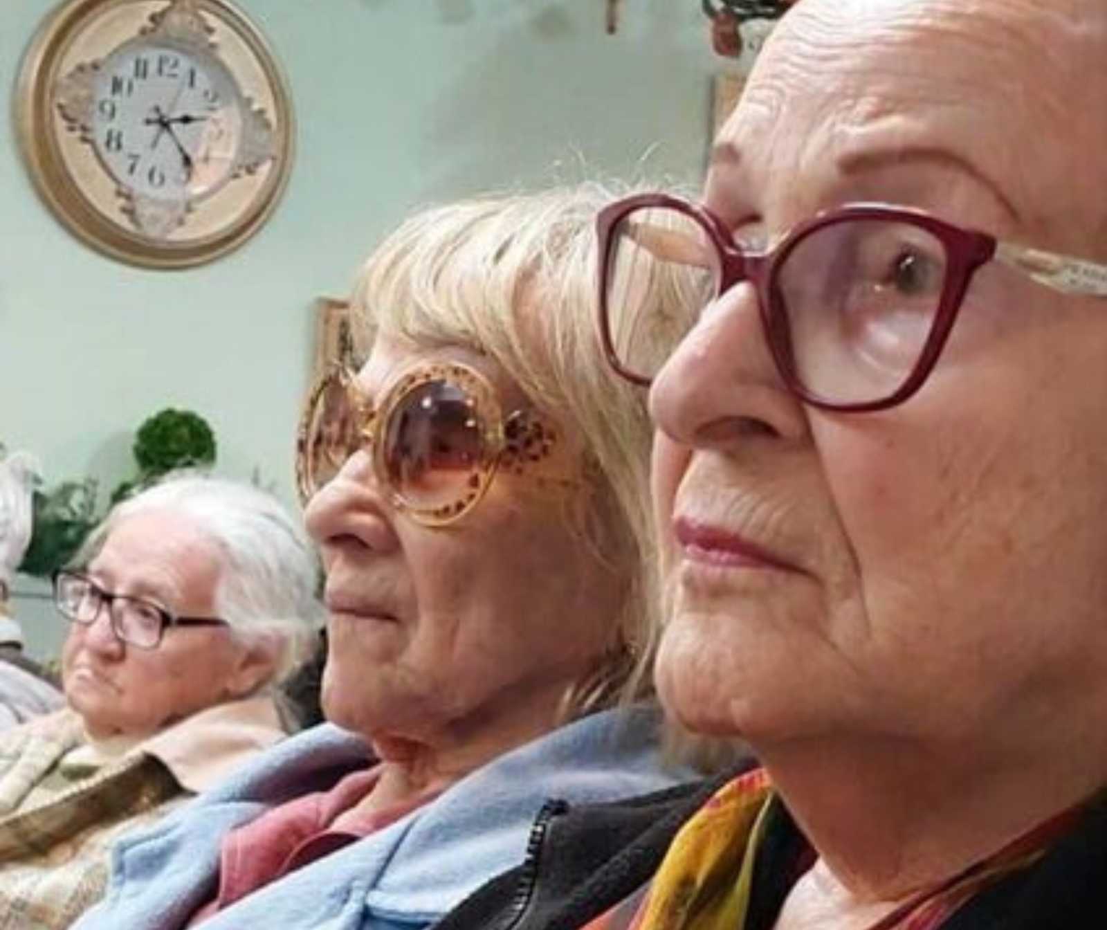 drei alte Damen nebeneinander schauen in die gleiche Richtung. An der Wand des Zimmers hängt eine Uhr; Pflanzen sind im Hintergrund zu sehen