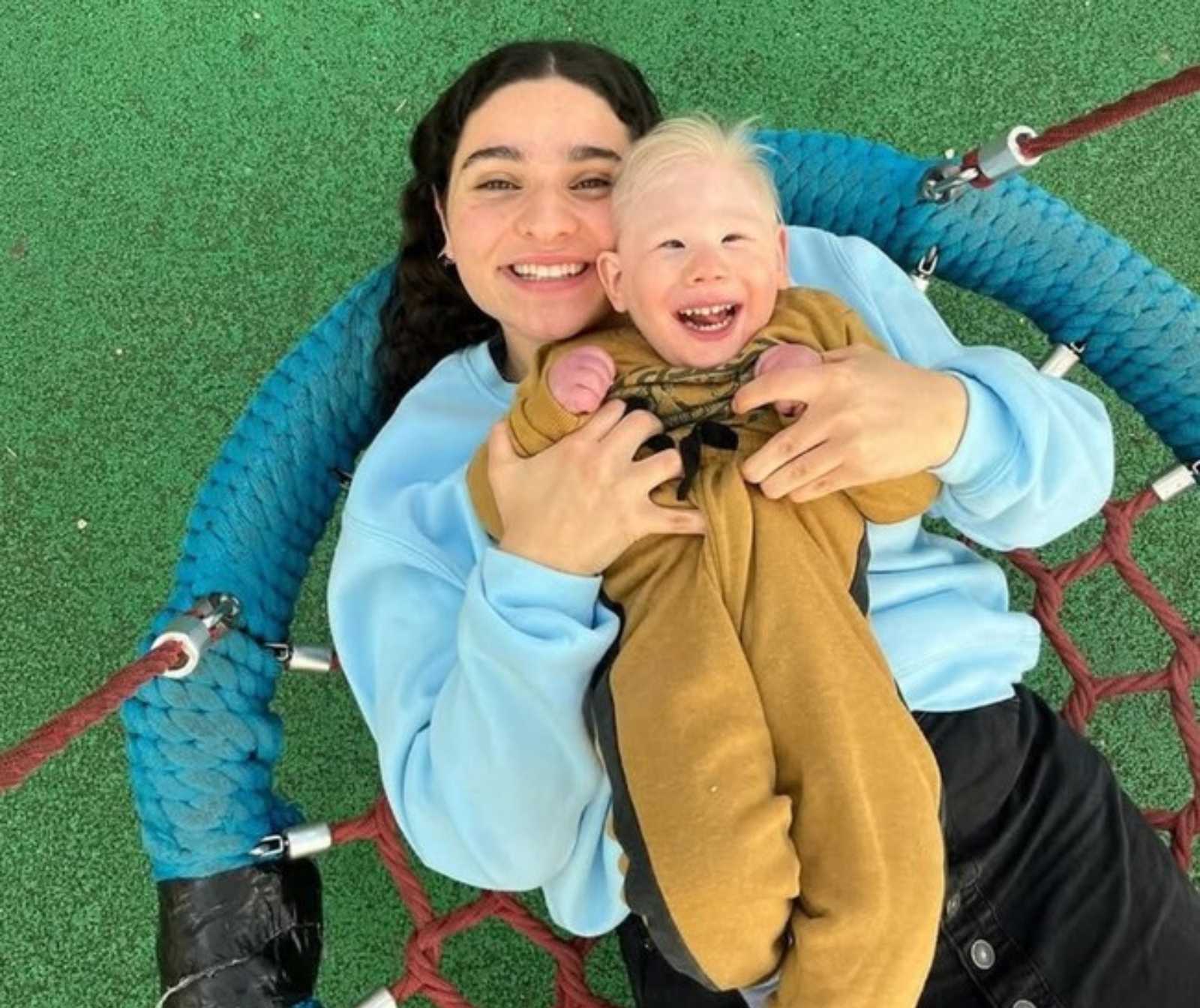 eine Aufnahme von oben auf eine Frau. die ein behindertes Kind auf ihrem Oberkörper liegen hat. Beide lächeln in die Kamera