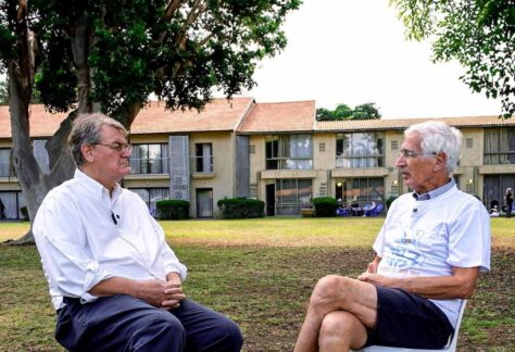zwei Männer sitzen sich auf Stühlen im Freien entgegen. Im Hintergrund ist ein Wohnkomples