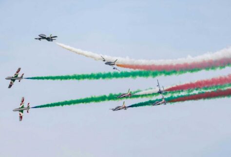 mehrere Kunstflugzeuge fliegen in Formation durch einen klaren Himmel und hinterlassen Rauchstreifen in den Farben Grün, Weiß und Rot.