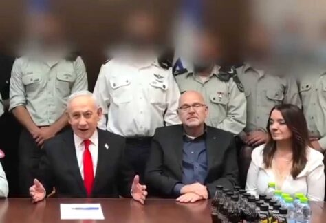 mehrere Personen sitzen und stehen um einen Tisch, während ein Mann im Anzug und roter Krawatte spricht und gestikuliert