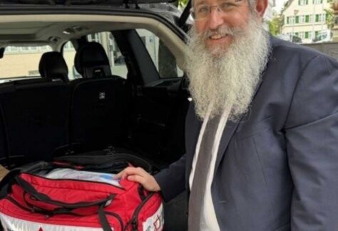 ein Rabbiner steht hinten am geöffneten Kofferraum und hält seine Hand an einer rot/weißen Tasche