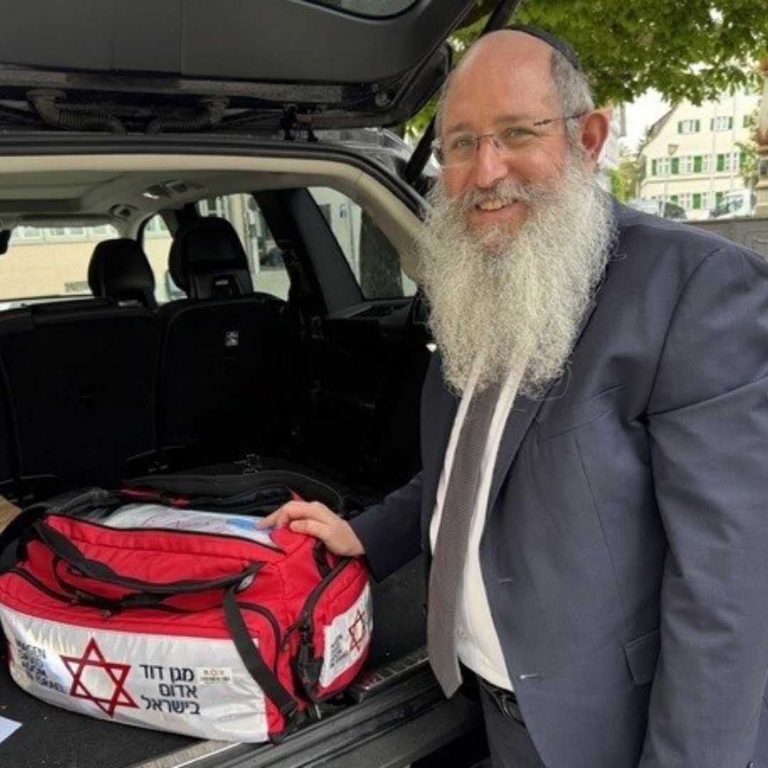 ein Rabbiner steht hinten am geöffneten Kofferraum und hält seine Hand an einer rot/weißen Tasche